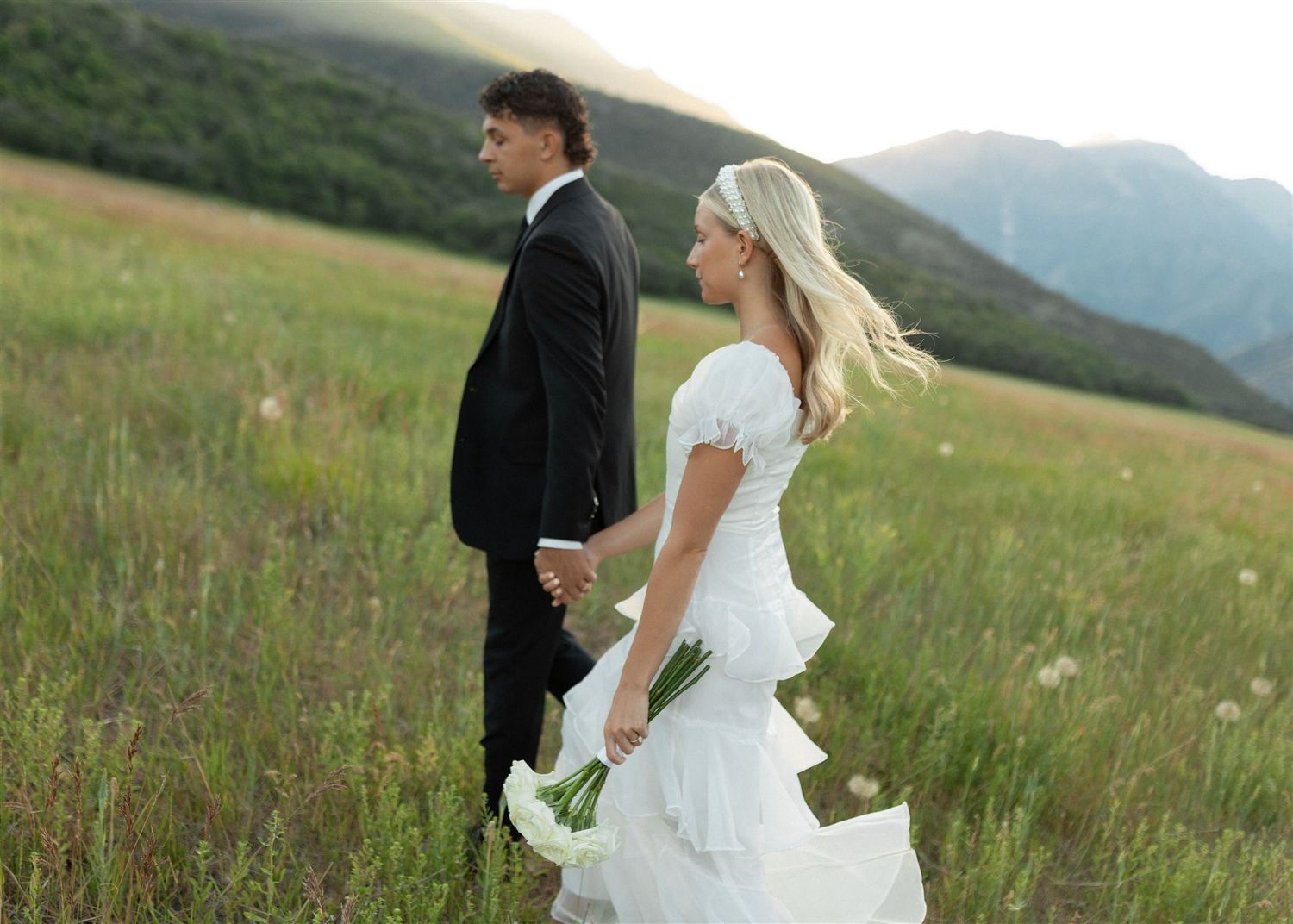 A bride and groom walking together through a green mountain meadow